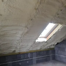 Poddasze z oknem dachowym po ociepleniu pianką, widoczne kołki mocujące i surowe ściany z bloczków betonowych.