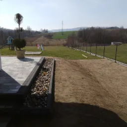 Widok na świeżo wyrównaną ziemię pod trawnik, z widocznym fragmentem tarasu z kamienną rabatą i nowym ogrodzeniem siatkowym w tle.