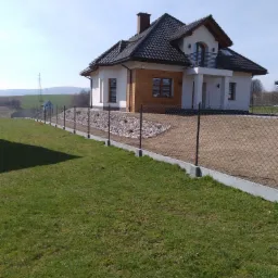 Nowoczesny dom jednorodzinny z brązowym dachem i drewnianą elewacją, otoczony siatką ogrodzeniową na betonowej podmur&oacute;wce i rabatą z białych kamieni, na pierwszym planie zielony trawnik.