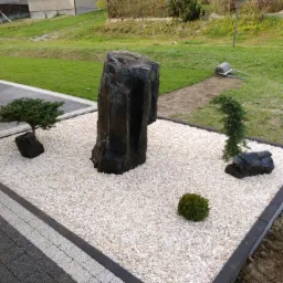 Ogród zen z dużym, ciemnym kamieniem centralnym, otoczony białym żwirem i niskimi roślinami iglastymi, obrzeże z ciemnych kostek.