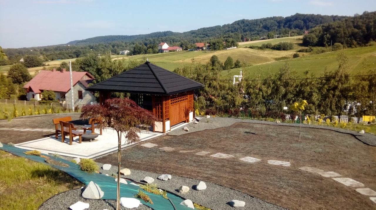 Widok z góry na zagospodarowany teren z altaną, drzewkiem japońskim, ścieżką z kamieni i przygotowanym podłożem pod trawnik na tle pagórkowatego krajobrazu.