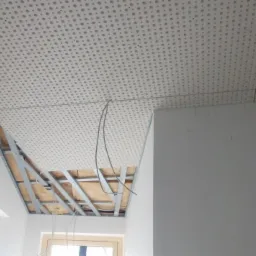 Montaż sufitu podwieszanego z płyty perforowanej, widoczna metalowa konstrukcja nośna i wystające kable, otwór wentylacyjny w ścianie, okno w tle.