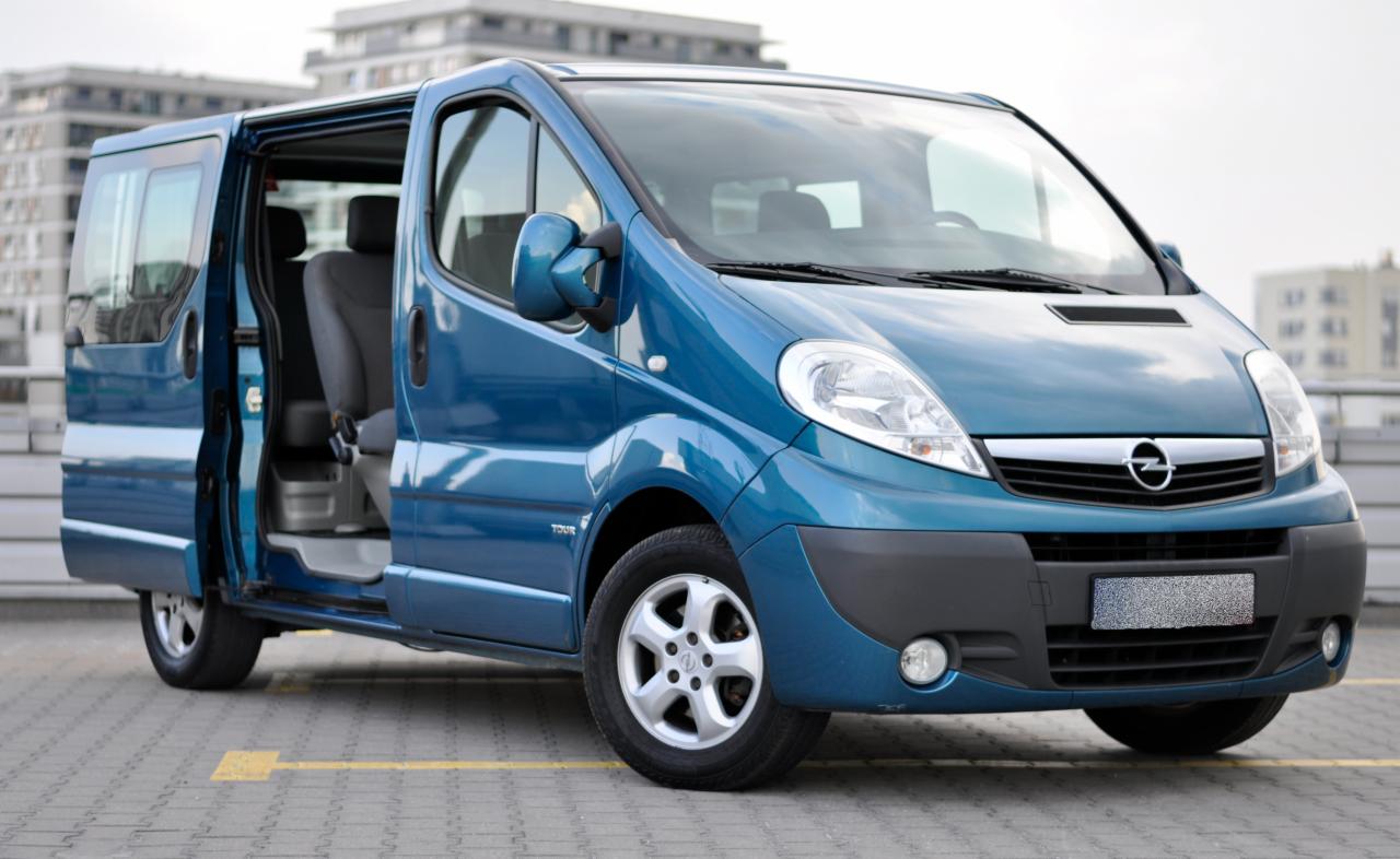 Niebieski Opel Vivaro Tour z otwartymi drzwiami przesuwnymi na tle nowoczesnego budynku.