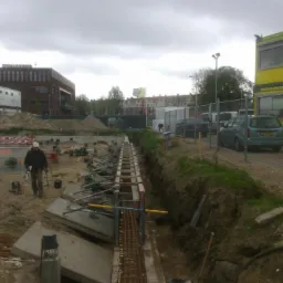 Budowa fundamentów: Wykop z szalunkiem i zbrojeniem, w tle widoczny pracownik w kasku, betonowe elementy i budynek biurowy.