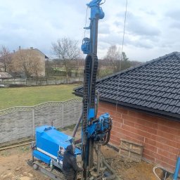Greenhouse-RS Sp. z o.o. - Niebieska wiertnica na gąsienicach, obsługiwana przez pracownika w ubłoconym ubraniu roboczym, wiercąca otwór w ziemi obok ceglanego budynku z ciemnym dachem, widok z góry.