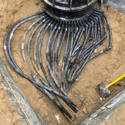 Greenhouse-RS Sp. z o.o. - Czarna, żebrowana studzienka z podłączonymi licznymi, czarnymi przewodami w wykopie otoczonym drewnianą szalunką, obok leży młotek brukarski z żółtym trzonkiem.