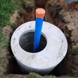 Greenhouse-RS Sp. z o.o. - Betonowy krąg studni w wykopie ziemnym z wystającą niebieską rurą zabezpieczoną pomarańczową taśmą, w tle fragment koparki.