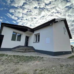 Nowo otynkowany dom jednorodzinny z ciemną podmurówką i schodami wejściowymi, widok z dołu na tle pochmurnego nieba.