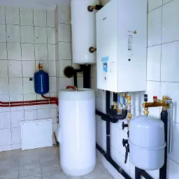 Zestaw urządzeń grzewczych w pomieszczeniu z białymi płytkami, widoczny biały zasobnik akumulacyjny, wiszący bojler oraz niebieski zbiornik wyrównawczy, połączone czarnymi rurami.