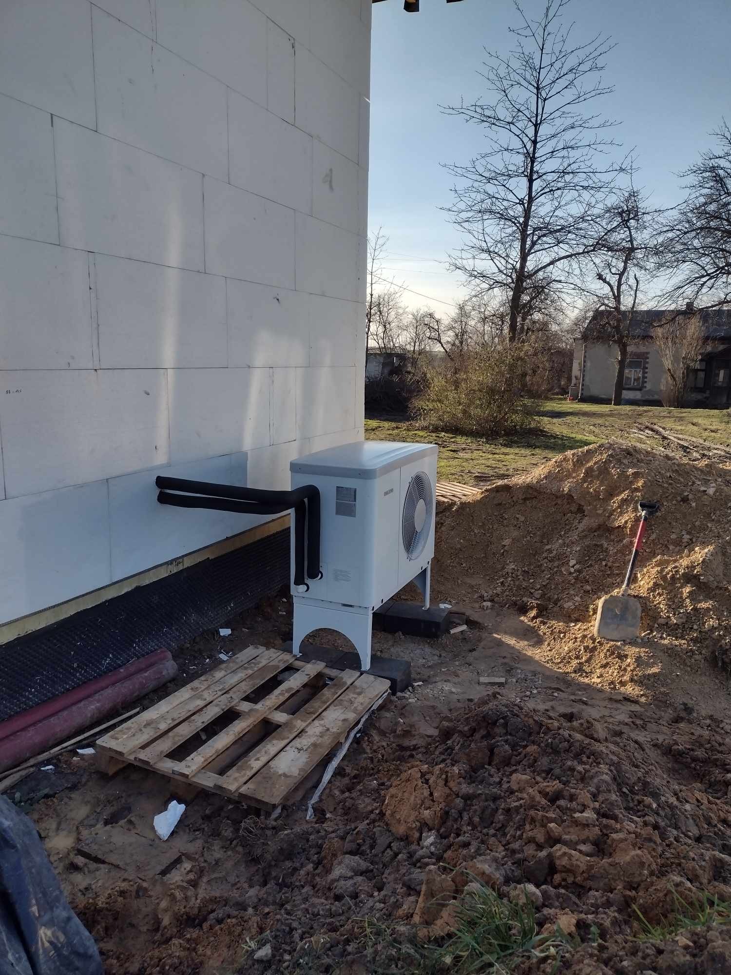 Biała pompa ciepła zamontowana przy ścianie domu z białych bloczków, widoczne czarne rury łączące urządzenie z wnętrzem, paleta drewniana na ziemi, w tle sterta ziemi i łopata.