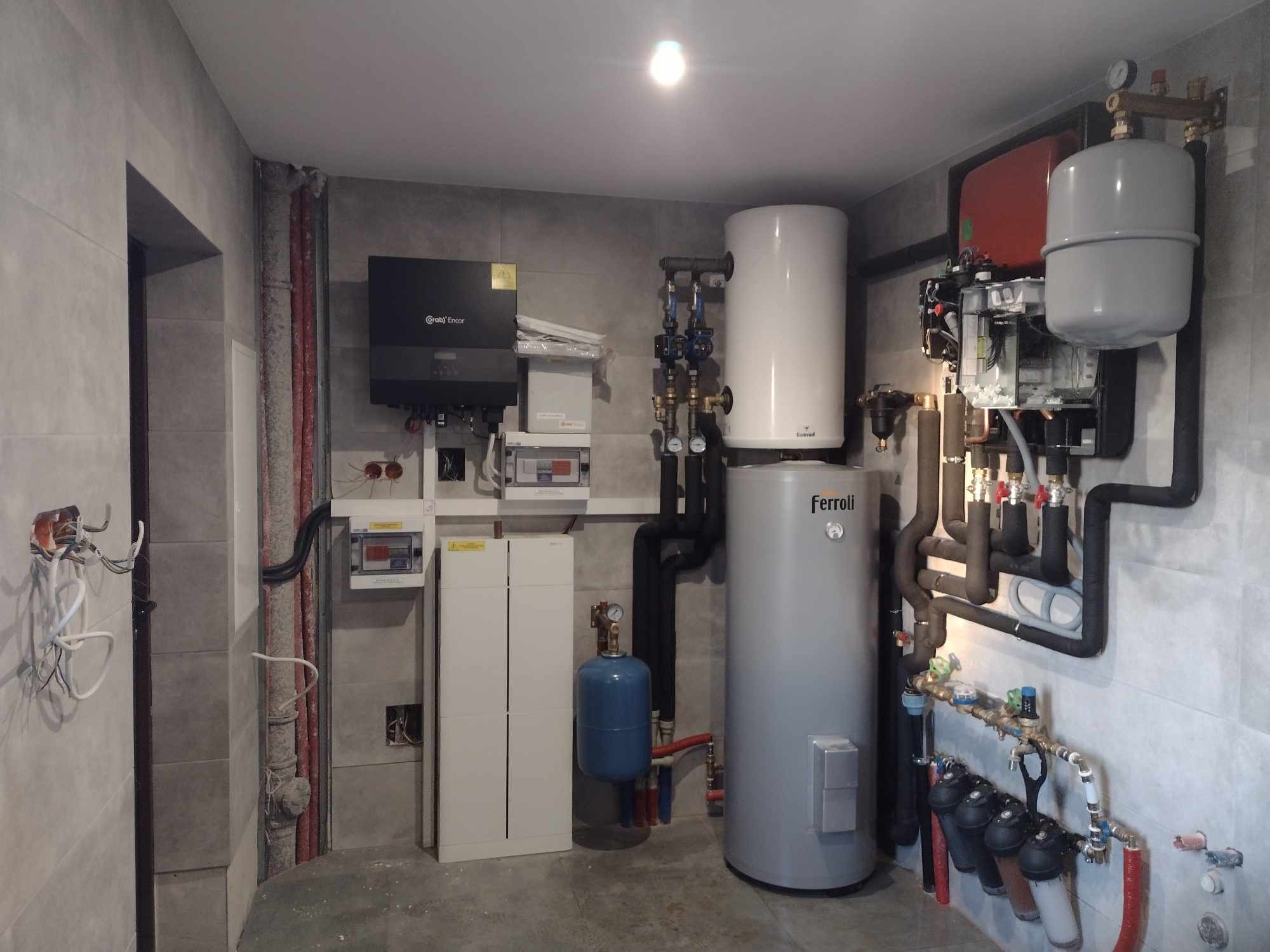 Zestaw urządzeń w kotłowni: zbiornik buforowy Ferroli, pompa ciepła powietrze-woda, filtry wody, rozdzielacze hydrauliczne i rury w pomieszczeniu wykończonym szarą płytką.