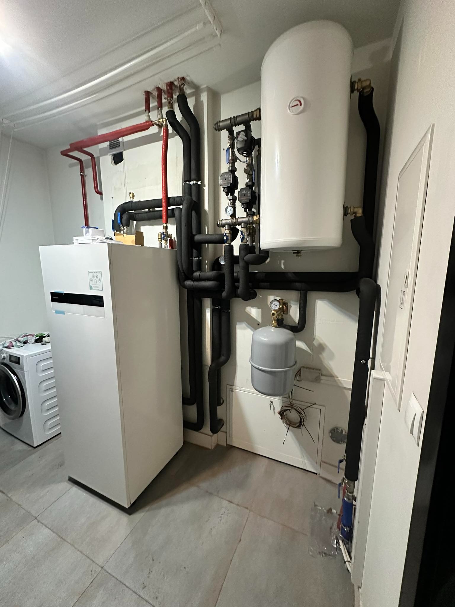 Wnętrze kotłowni z zainstalowaną białą pompą ciepła Panasonic, zbiornikiem na wodę i szarym naczyniem przeponowym; widoczne rury w izolacji oraz pralka w tle.