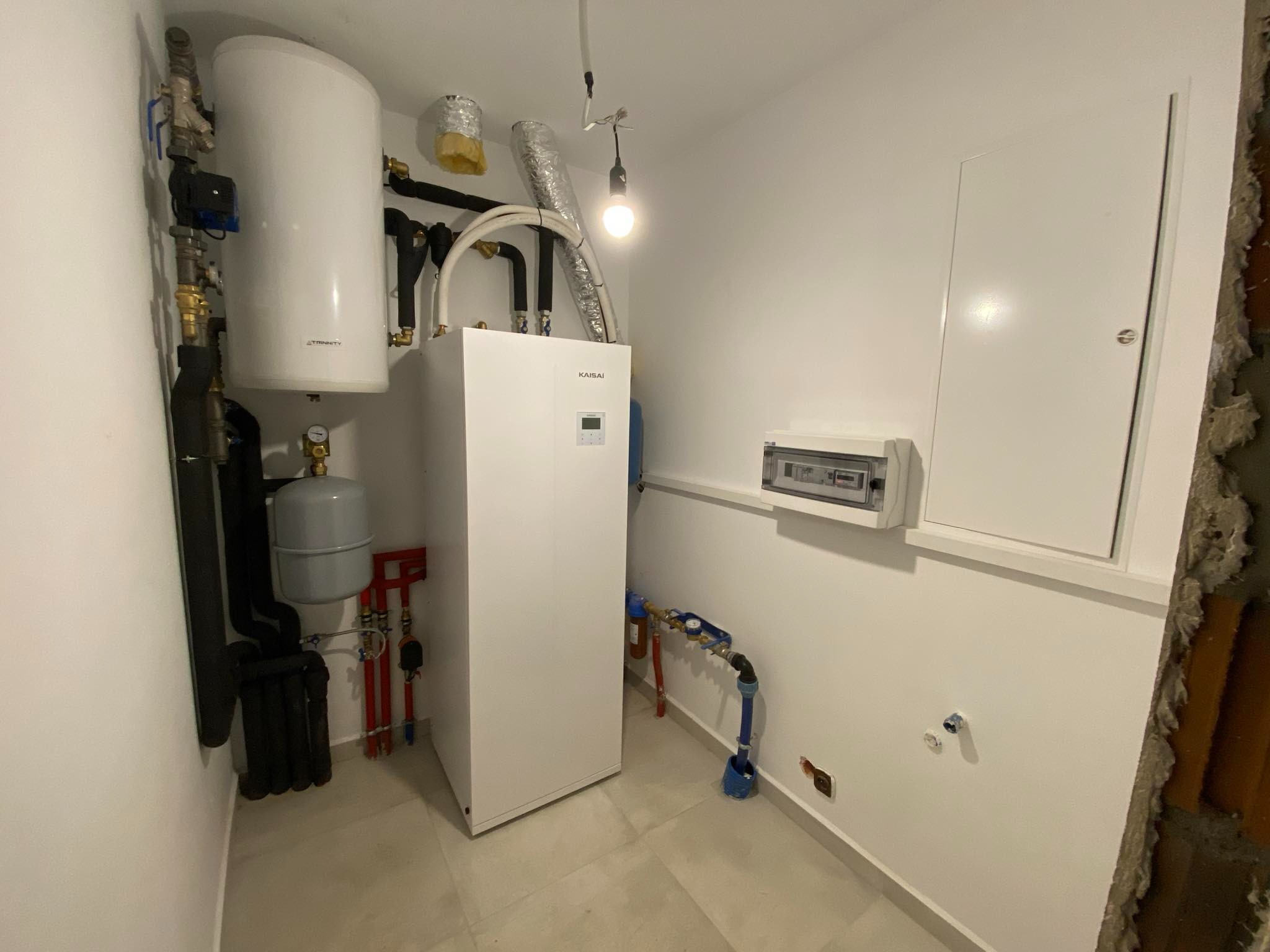 Biała pompa ciepła Kaisai w kotłowni, z widocznymi rurami, zbiornikiem Trinity, skrzynką elektryczną i surowym wykończeniem ściany po prawej stronie.