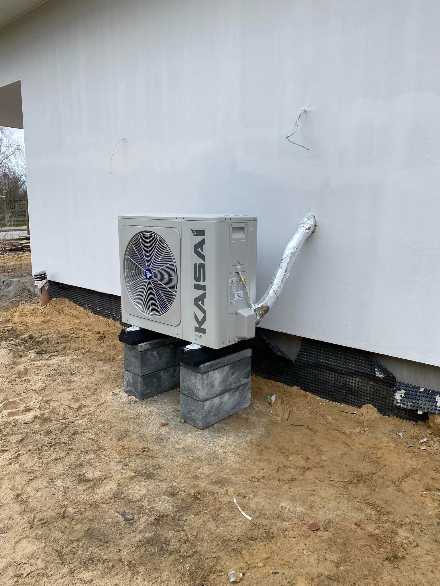 Zewnętrzna jednostka klimatyzacji Kaisai zamontowana na betonowych blokach przy ścianie budynku w trakcie prac ziemnych.