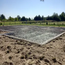 Zbrojenie płyty fundamentowej ułożone w szalunku na piasku, widok z góry, w tle ogrodzenie z siatki i drzewa.