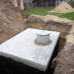 Betonowy zbiornik szamba z pokrywą umieszczony w wykopie, z widoczną izolacją przeciwwodną, na tle wiejskich zabudowań i sterty ziemi.