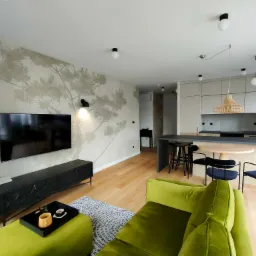 Salon z zieloną sofą i ścianą z motywem drzewa. Nowoczesne oświetlenie i drewniana podłoga dodają wnętrzu ciepła i elegancji. Elementy dekoracyjne tworzą spójną całość.