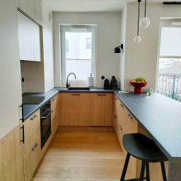 SPACE MANUFACTURE - Jasna kuchnia z drewnianymi frontami szafek, czarnym blatem i minimalistycznym oświetleniem. Na blacie czerwona misa z owocami i czajnik. Widok z perspektywy baru.