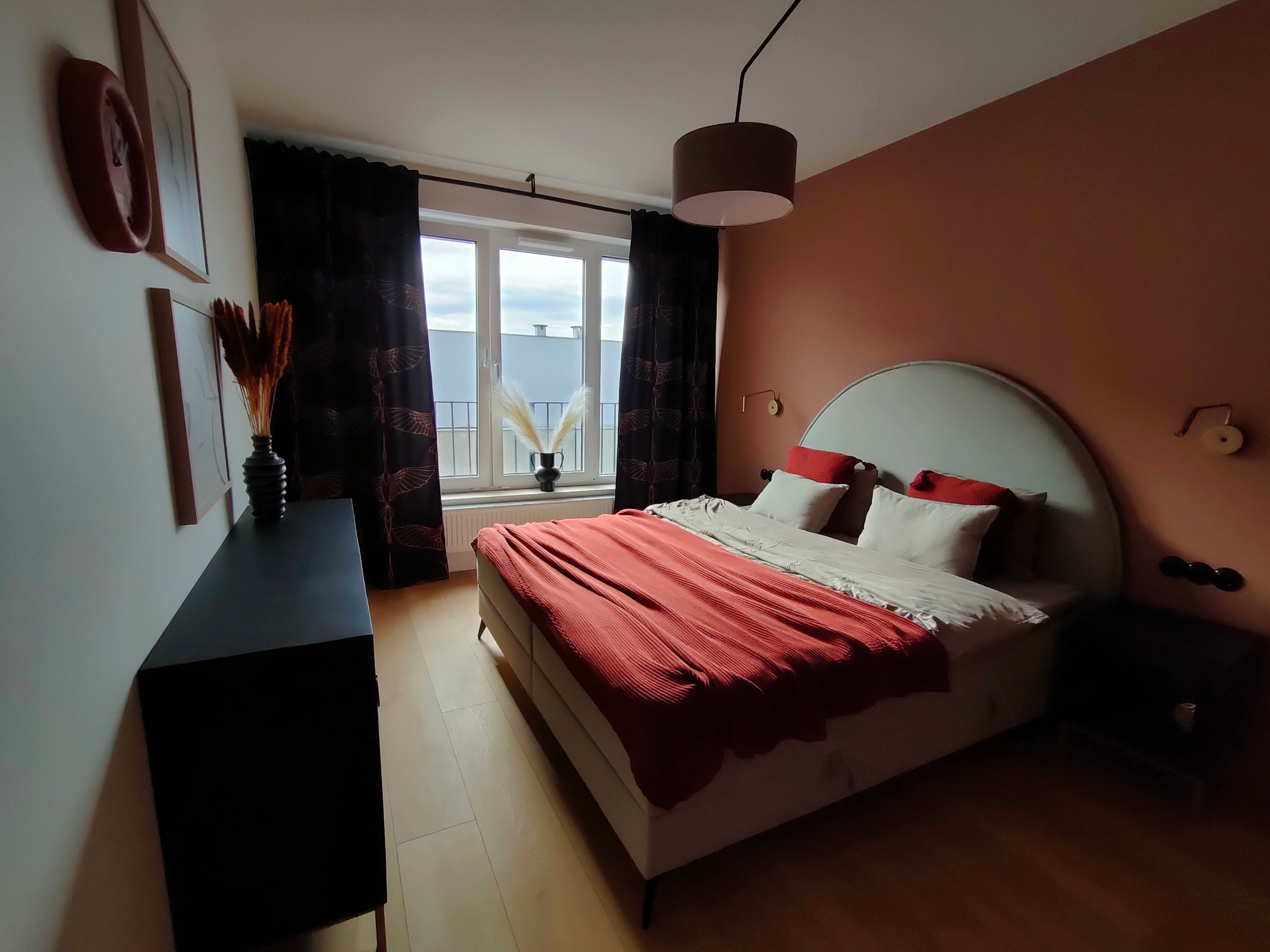 Sypialnia z łóżkiem, czerwoną narzutą i czarną komodą. Okno z ciemnymi zasłonami, widok na balkon. Ściana w kolorze terakoty, lampka nocna i dekoracje.