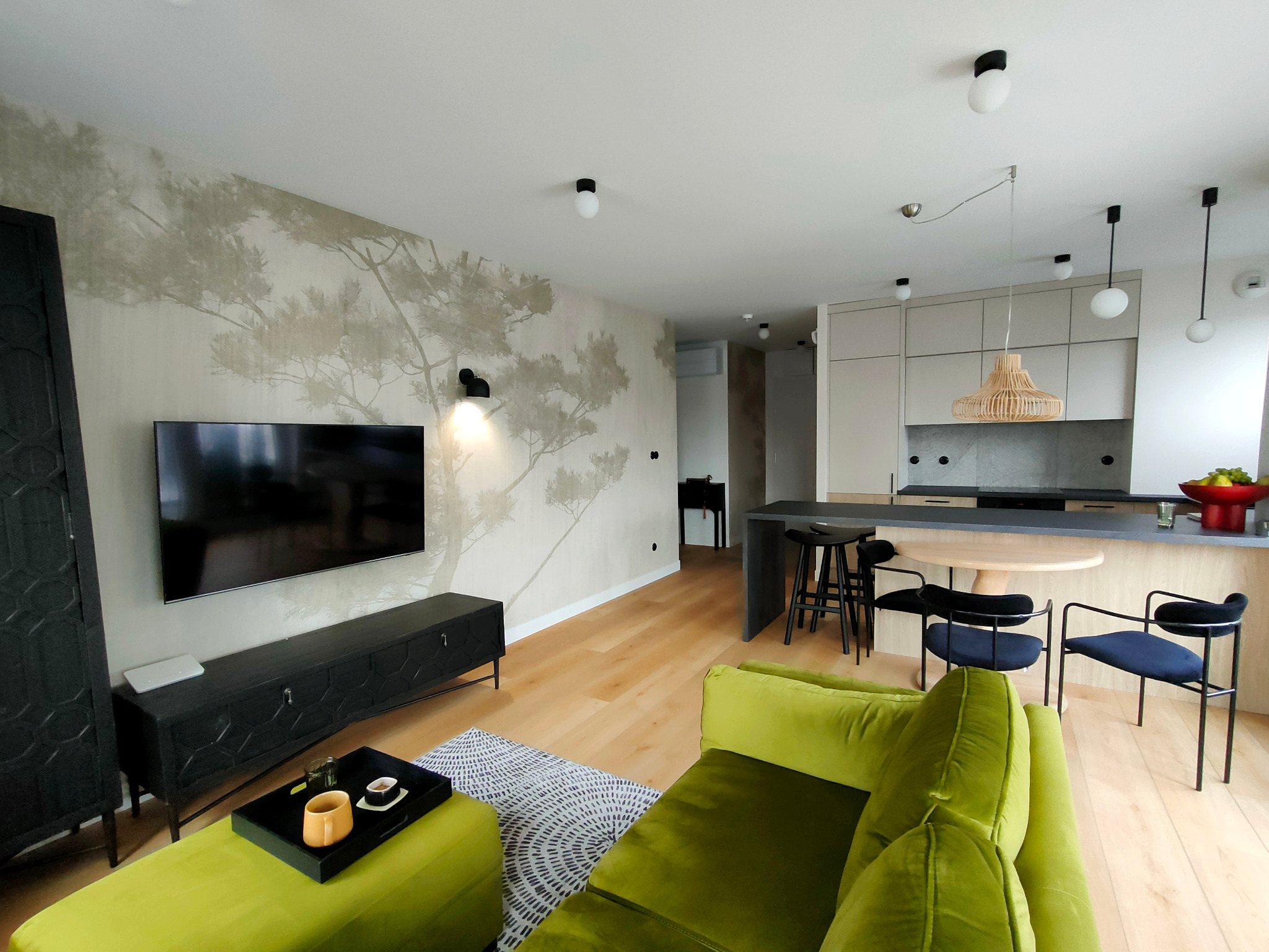 Salon z zieloną sofą i ścianą z motywem drzewa. Nowoczesne oświetlenie i drewniana podłoga dodają wnętrzu ciepła i elegancji. Elementy dekoracyjne tworzą spójną całość.