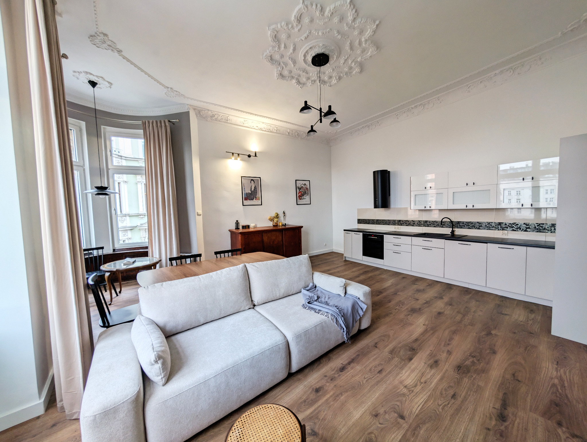 Jasne, przestronne wnętrze z sofą, stołem i aneksem kuchennym. Dekoracyjne sztukaterie sufitowe i drewniana podłoga dodają elegancji. Widok na budynek zza okna.