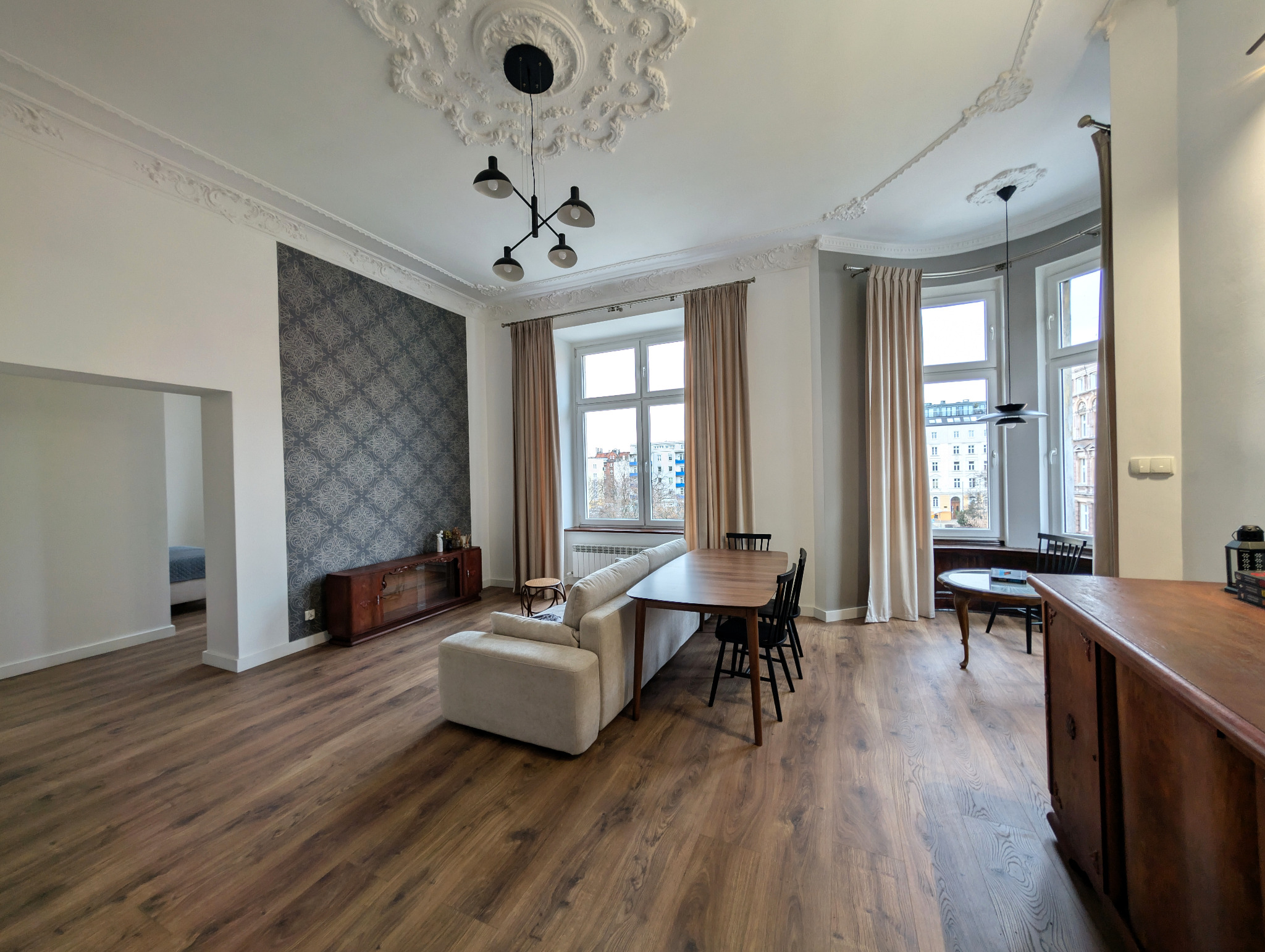 Przestronne wnętrze mieszkania z drewnianą podłogą, sofą, stołem z krzesłami i ozdobnymi sztukateriami sufitowymi. Widok na okno z beżowymi zasłonami.