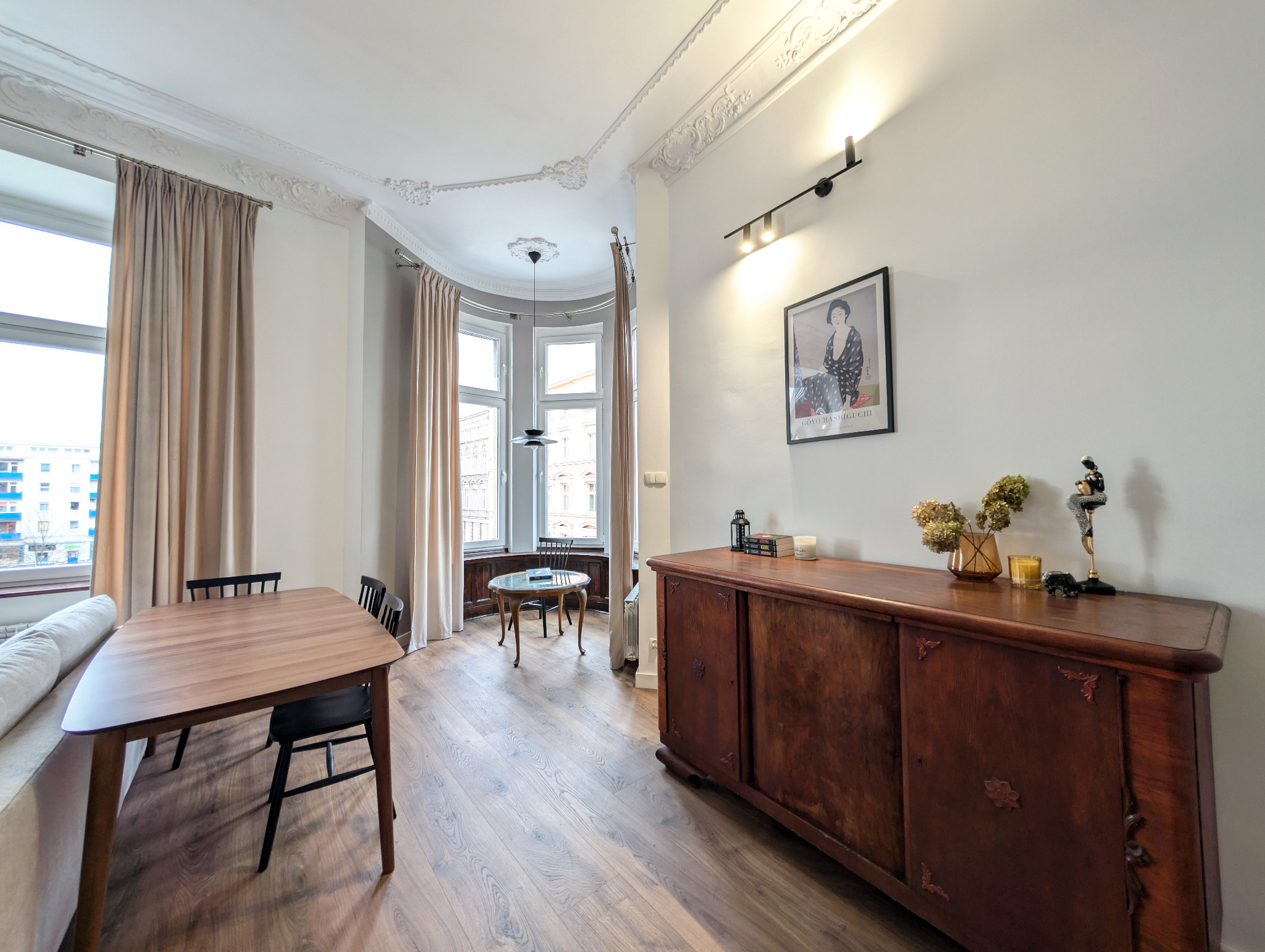 Stylowa aranżacja salonu we Wrocławiu z drewnianym kredensem, okrągłym stolikiem w wykuszowym oknie i stołem jadalnym. Klasyczne detale sufitowe i nowoczesne oświetlenie.