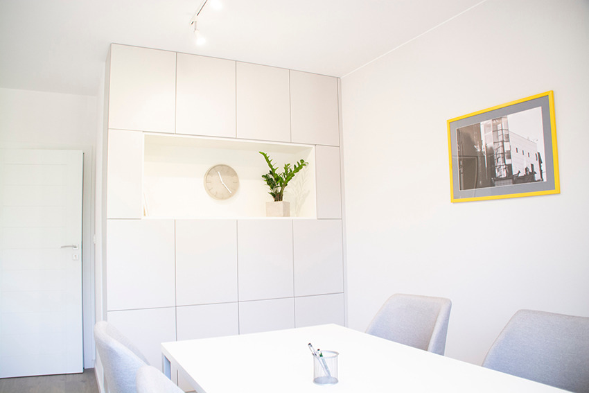 Białe, minimalistyczne wnętrze biura z wbudowanymi szafkami, zegarem, rośliną i obrazem w żółtej ramie. Stół i krzesła w jasnych odcieniach. Nowoczesny design.