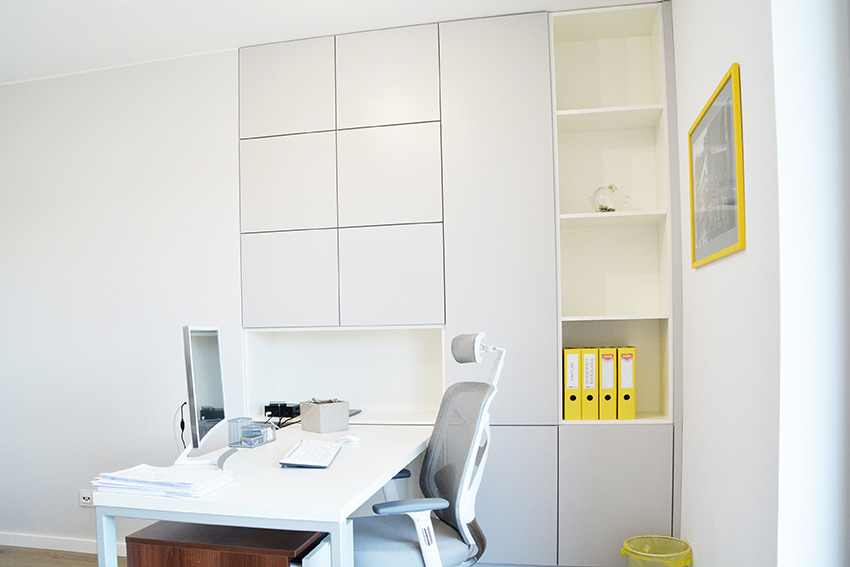 Nowoczesne biuro: białe meble w zabudowie, ergonomiczne krzesło, minimalistyczny design, żółte segregatory i rama obrazu dodają akcentu kolorystycznego.