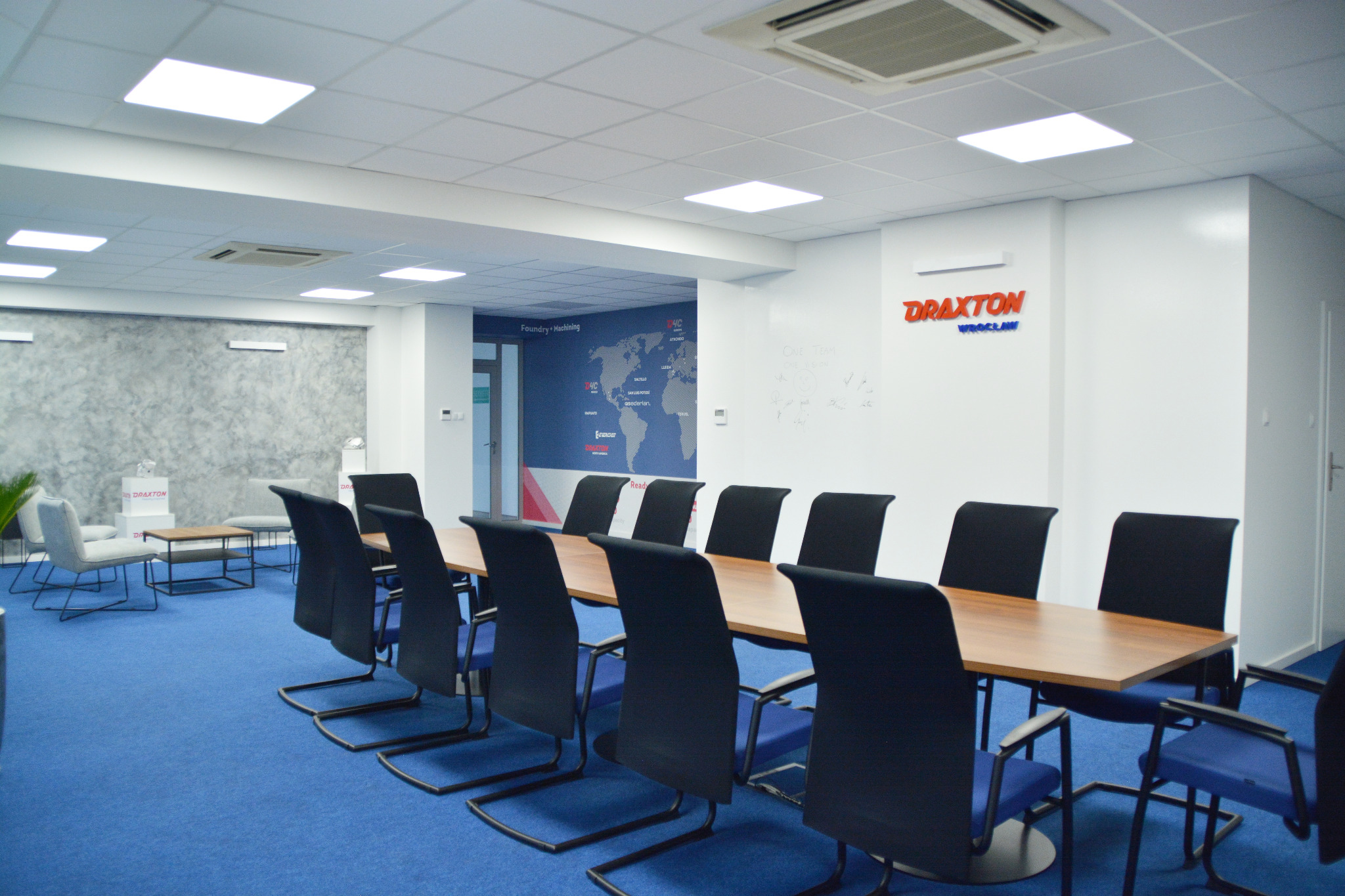 Jasne wnętrze biura z długim stołem konferencyjnym, niebieską wykładziną i białymi ścianami. W tle logo firmy Draxton i mapa świata.