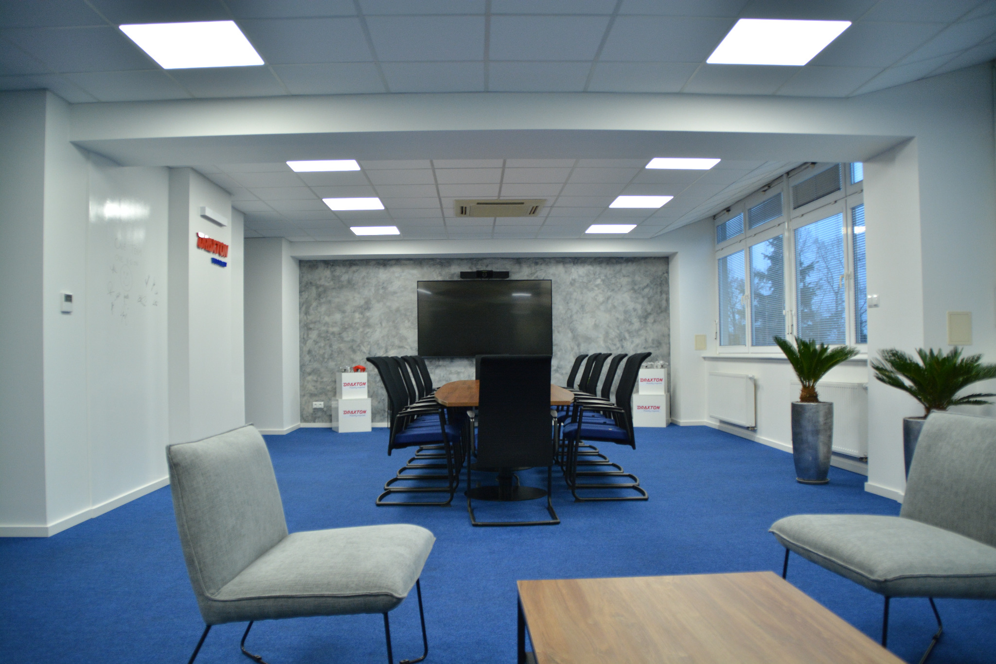 Nowoczesna sala konferencyjna z drewnianym stołem, czarnymi krzesłami i niebieską wykładziną. Widoczne rośliny w srebrnych donicach i ściana z teksturą betonu.