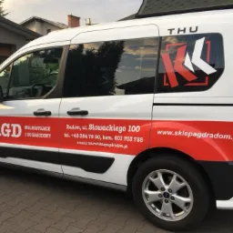 Biały bus z czerwonymi elementami graficznymi, logo firmy AGD, numerem telefonu i adresem strony internetowej sklepu z Radomia, zaparkowany na zewnątrz budynku.