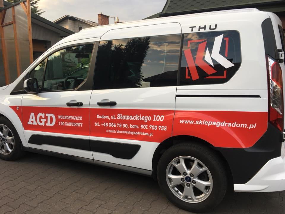 Biały bus z czerwonymi elementami graficznymi, logo firmy AGD, numerem telefonu i adresem strony internetowej sklepu z Radomia, zaparkowany na zewnątrz budynku.