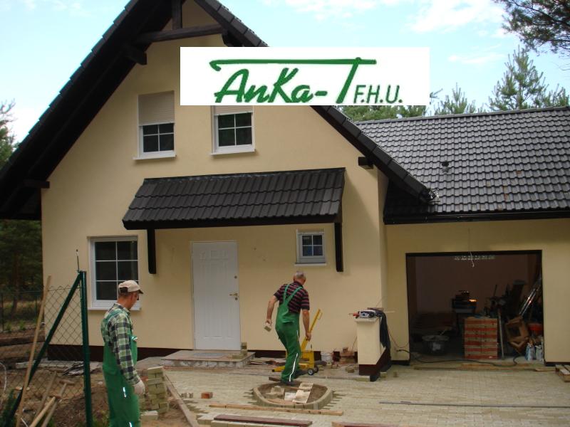 Dwóch pracowników w zielonych kombinezonach układa kostkę brukową przed nowo wybudowanym domem z garażem, widoczne narzędzia i materiały budowlane.