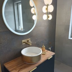 PRO-CONSTRUCTOR - Nowoczesna łazienka z okrągłym lustrem LED, złotą armaturą i umywalką. Ciemne płytki i drewniany blat szafki dodają elegancji. Stylowa lampa sufitowa.
