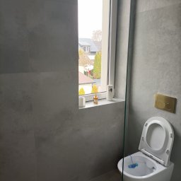 PRO-CONSTRUCTOR - Szara łazienka z podwieszaną toaletą i oknem z widokiem na domy. Minimalistyczny design, płytki imitujące beton, złoty przycisk spłuczki.