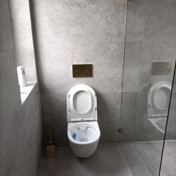 PRO-CONSTRUCTOR - Nowoczesna łazienka z szarymi płytkami, wiszącą toaletą z otwartą klapą i złotymi akcentami: przycisk spłuczki i szczotka do WC. Kabina prysznicowa ze szkła.