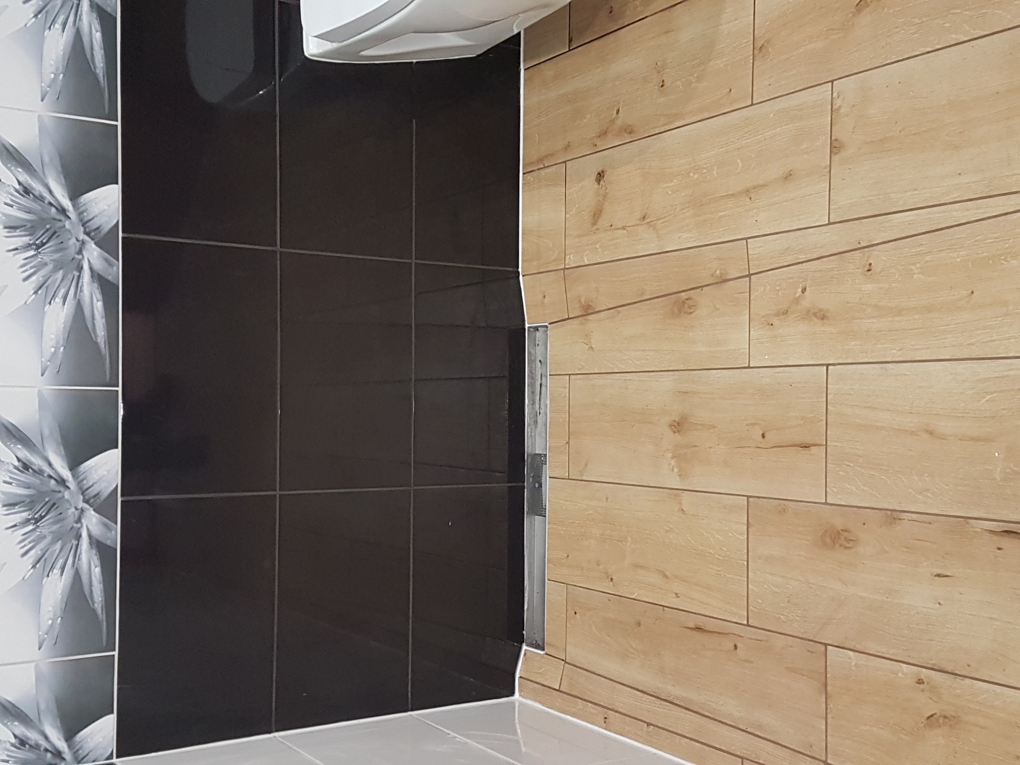 Fragment łazienki z ułożonymi płytkami: czarne, błyszczące płytki kwadratowe, płytki imitujące drewno ułożone poziomo oraz płytki z motywem kwiatowym w odcieniach szarości, widoczny fragment toalety.