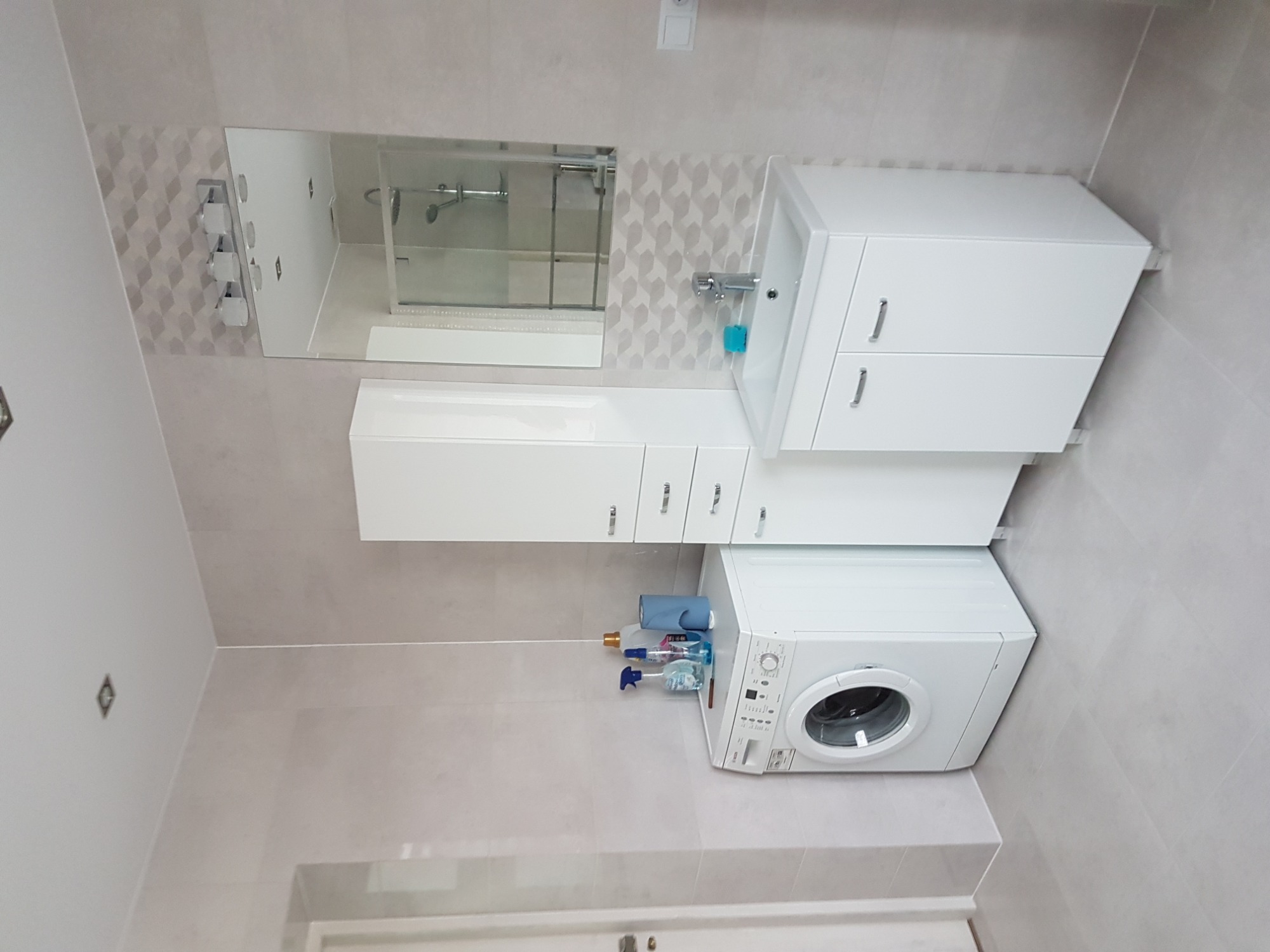 Jasna łazienka z pralką, umywalką z szafką, lustrem i wiszącymi szafkami, wykończona płytkami w odcieniach szarości, z widocznymi środkami czystości obok pralki.