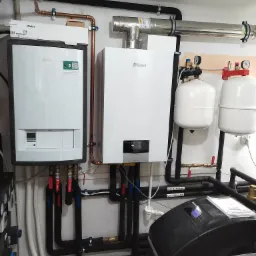 Dwa kotły gazowe marki Vaillant zamontowane na ścianie, z widocznymi rurami miedzianymi i czarnymi rurami izolowanymi, zbiorniki wyrównawcze oraz stacja uzdatniania wody Viteco Smart Flash.