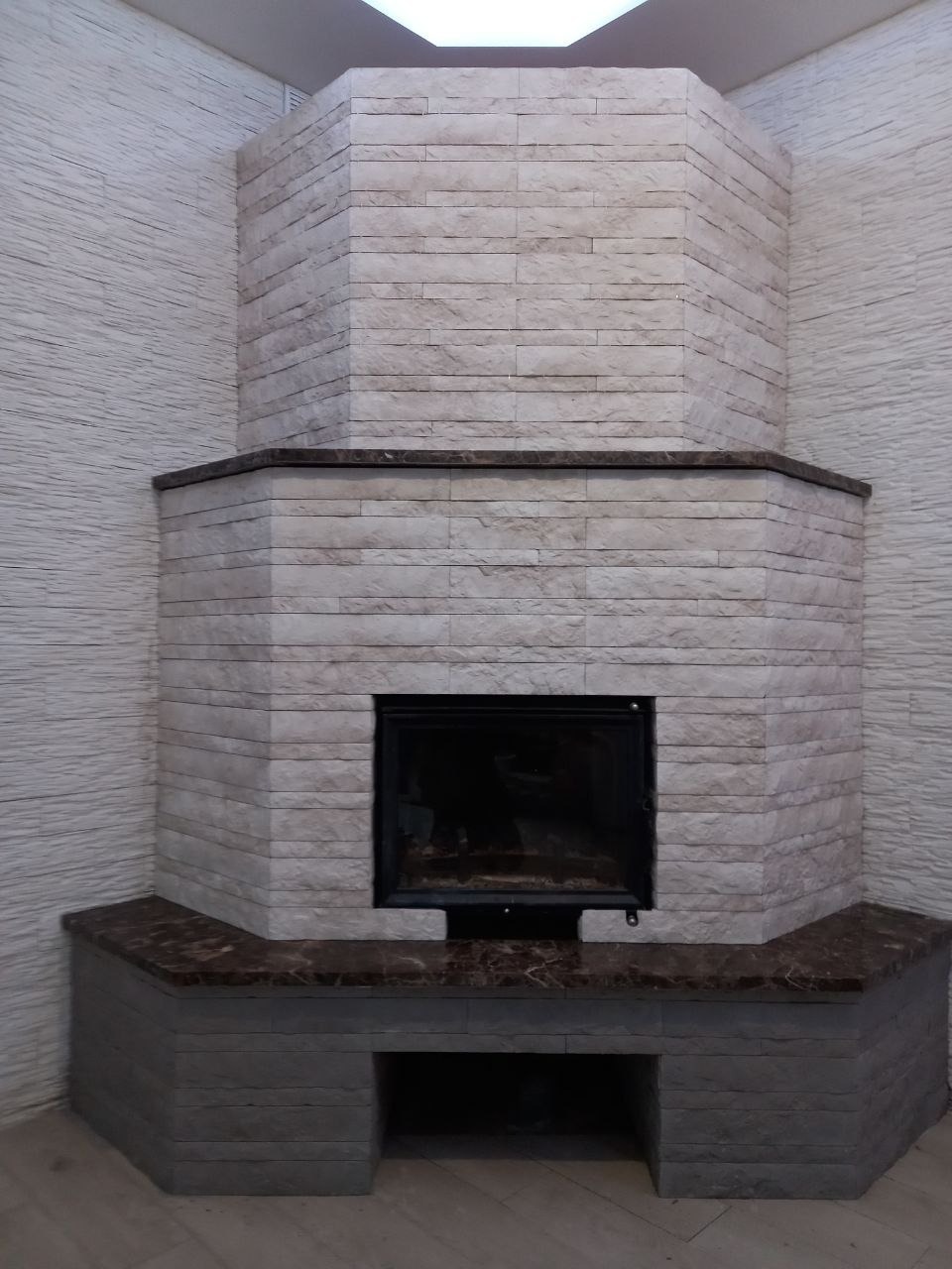 Wykończony kominek narożny z jasnych, prostokątnych płytek imitujących kamień, z ciemnym blatem i wkładem kominkowym. Ściana z fakturą.