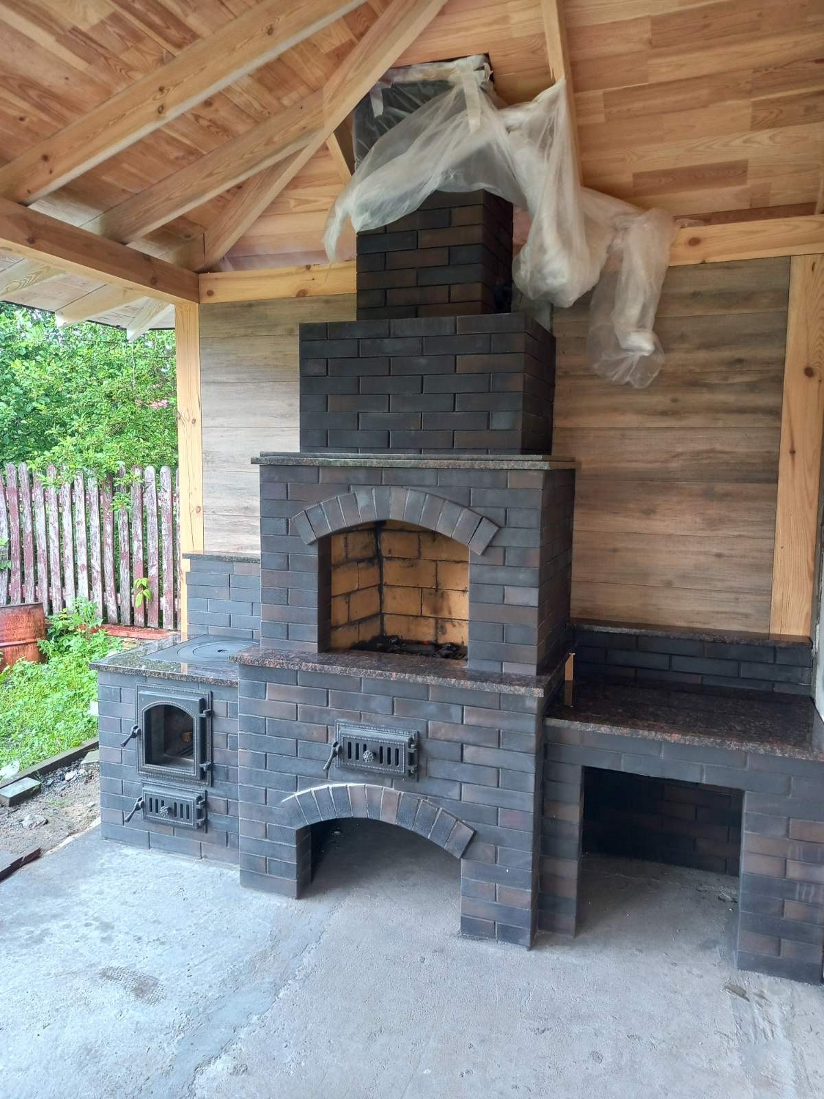 Zewnętrzny piec z ciemnej cegły z paleniskiem, okapem i blatem roboczym pod zadaszeniem. Widoczne detale wykończenia z granitu.