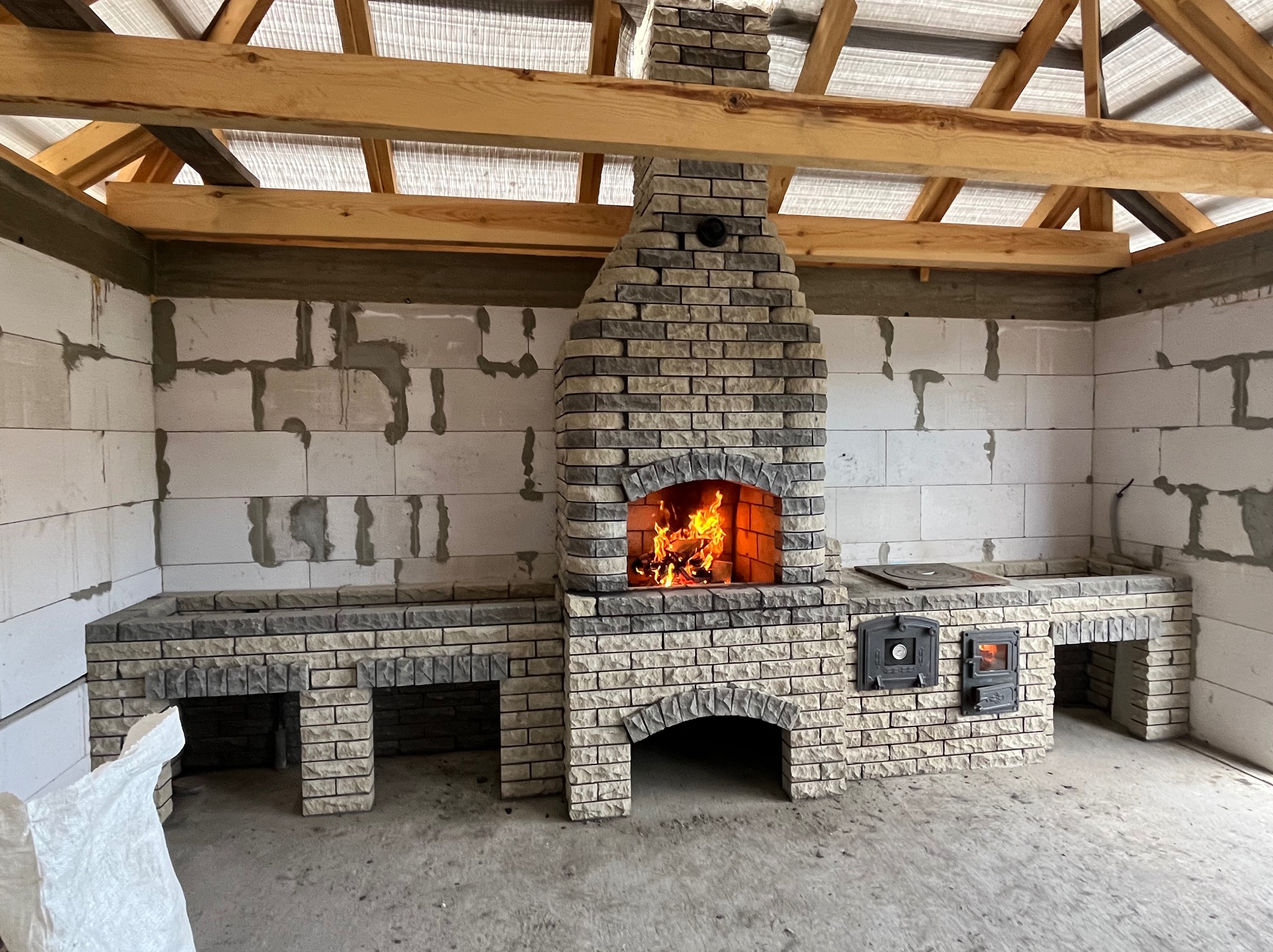 Murowany grill z cegły szamotowej w trakcie budowy, z paleniskiem i kominem, w surowym wnętrzu z widoczną konstrukcją dachu. Otwarty ogień w palenisku.