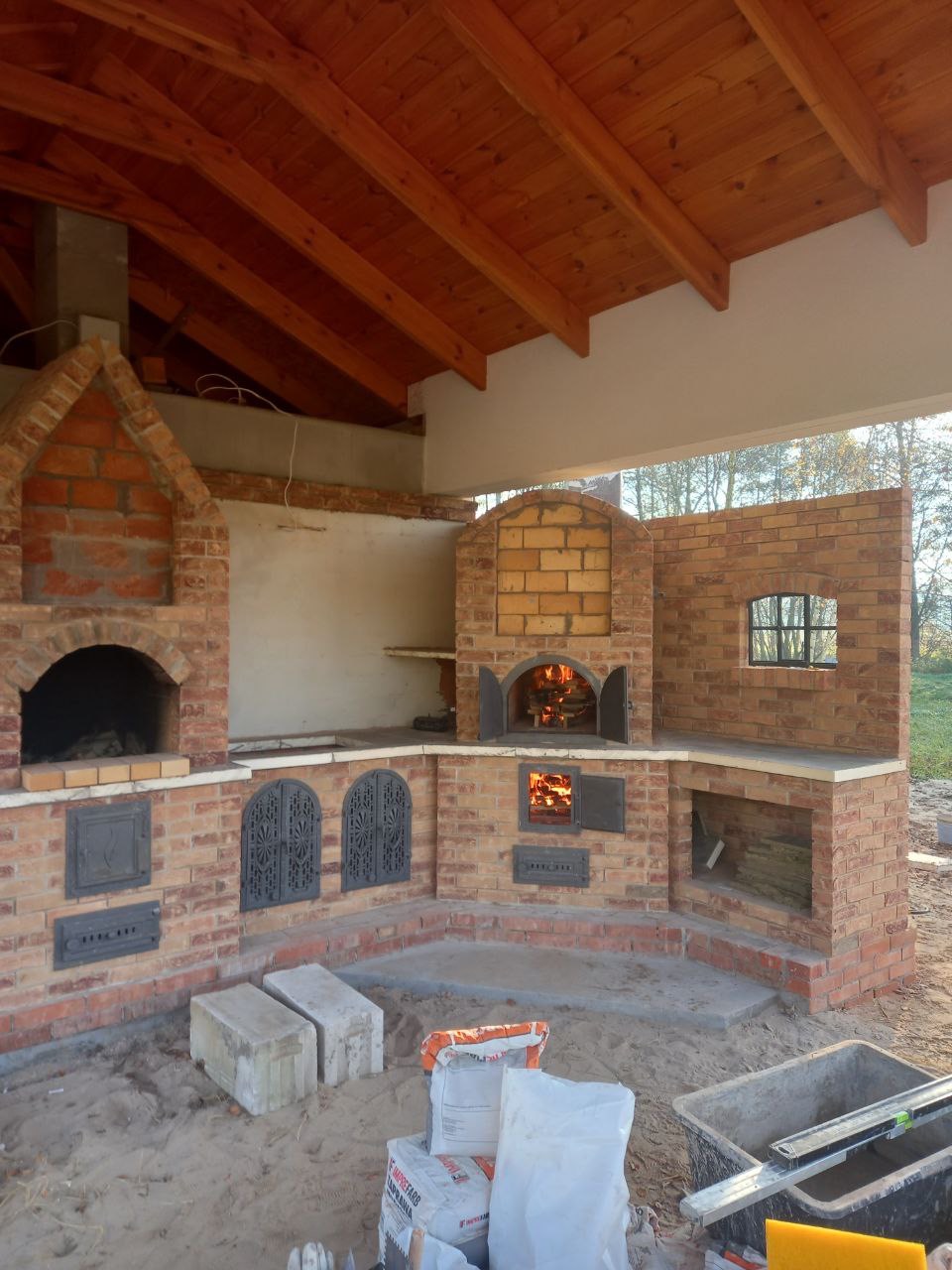 Zewnętrzna kuchnia z cegły z piecem chlebowym i grillem pod drewnianą wiatą. Widoczne detale wykończenia i palenisko z płomieniami. W tle zieleń.