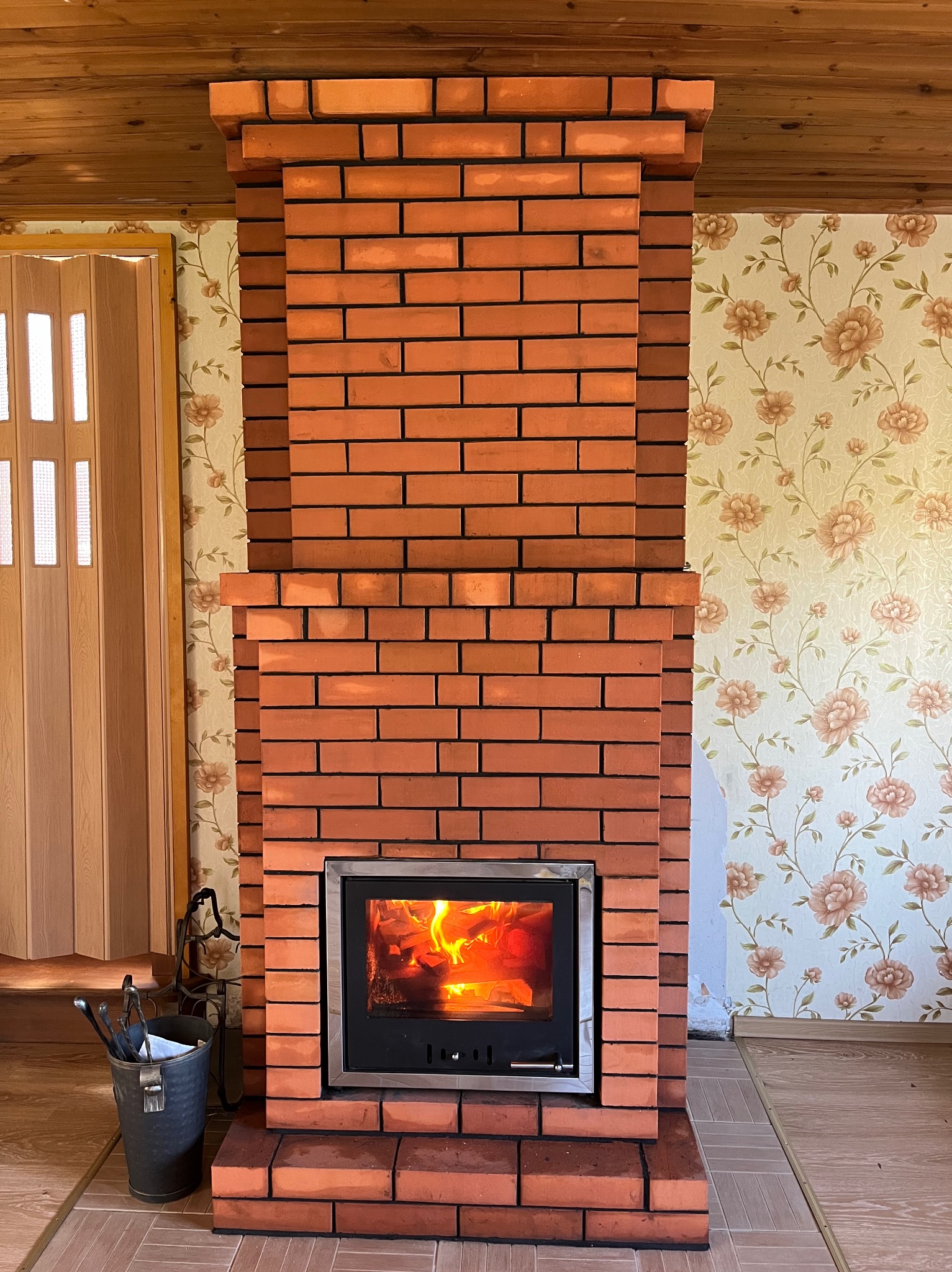 Murowany z cegły kominek z paleniskiem, widoczne płomienie. Obok drzwi harmonijkowe i wiadro z narzędziami kominkowymi. W tle ściana w kwiaty i drewniany sufit.