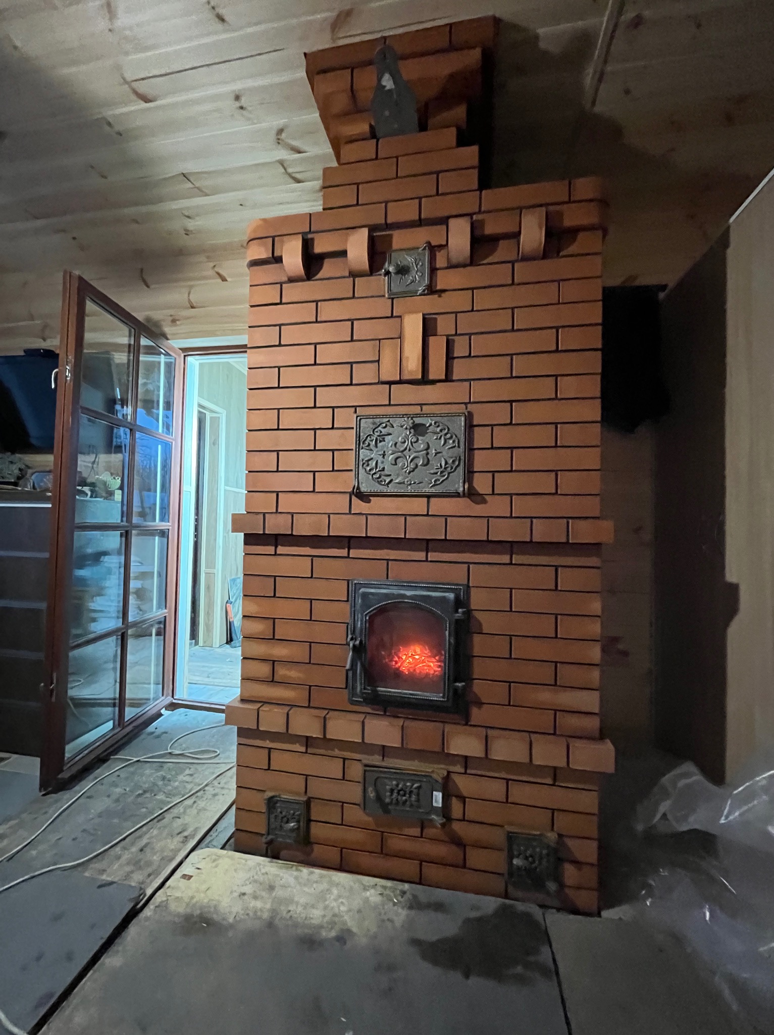Murowany piec z czerwonej cegły z żeliwnymi zdobieniami i palącym się ogniem w palenisku, widoczny w drewnianym wnętrzu domu z otwartym oknem.