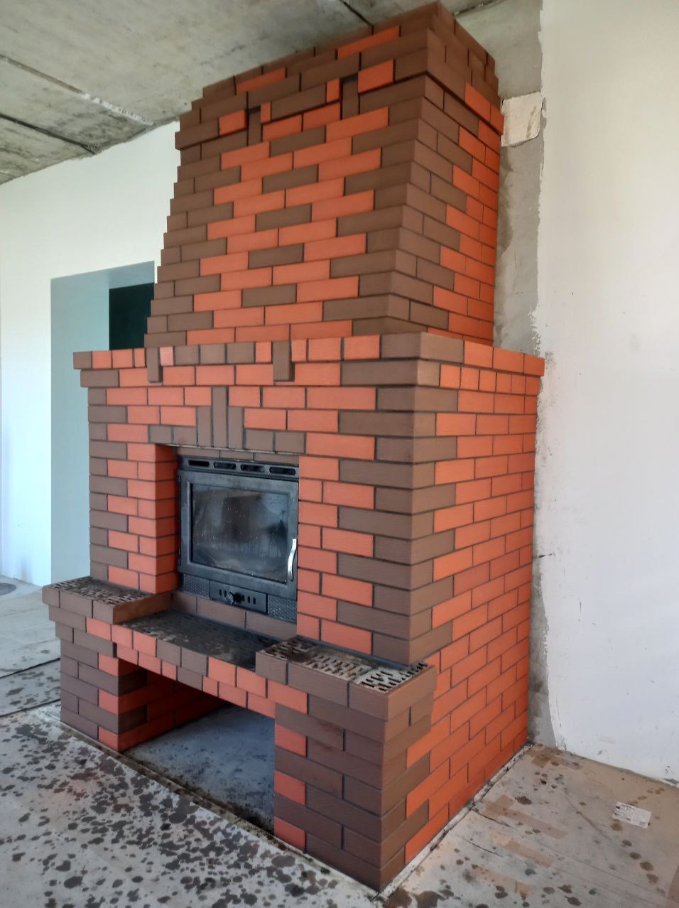 Nowoczesny kominek z cegły klinkierowej w odcieniach brązu i czerwieni, z czarnym wkładem kominkowym. Widoczne detale wykończenia i surowy stan pomieszczenia.