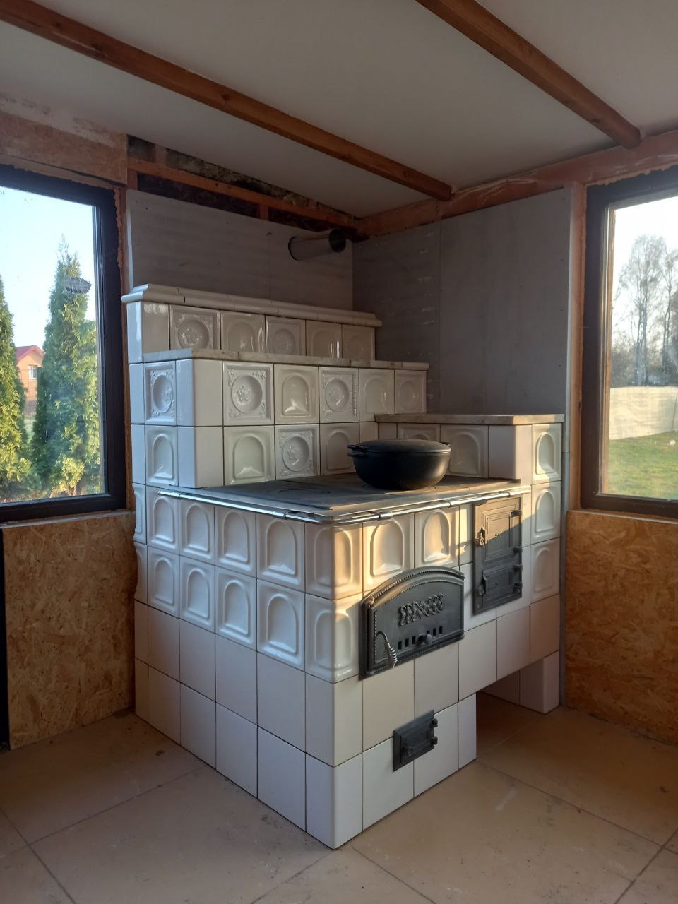 Biały, kaflowy piec z czarnym garnkiem na górze, w surowym wnętrzu z oknami. Widoczne drewniane elementy konstrukcyjne i płyty OSB.