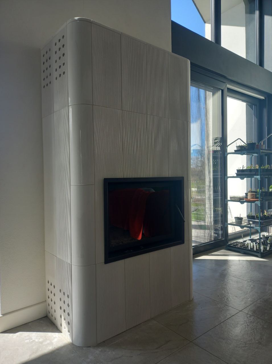 Biały, nowoczesny piec kaflowy z czarnym wkładem kominkowym, wykończony płytkami z delikatnym wzorem, na tle jasnego wnętrza z dużym oknem.