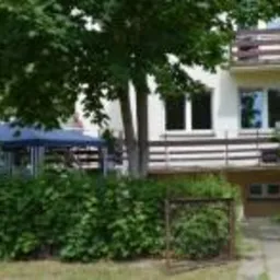 Dwupiętrowy budynek z balkonem, widoczny zza zielonych krzewów. Na balkonie biała balustrada. Z lewej strony widoczny fragment zadaszonego tarasu.